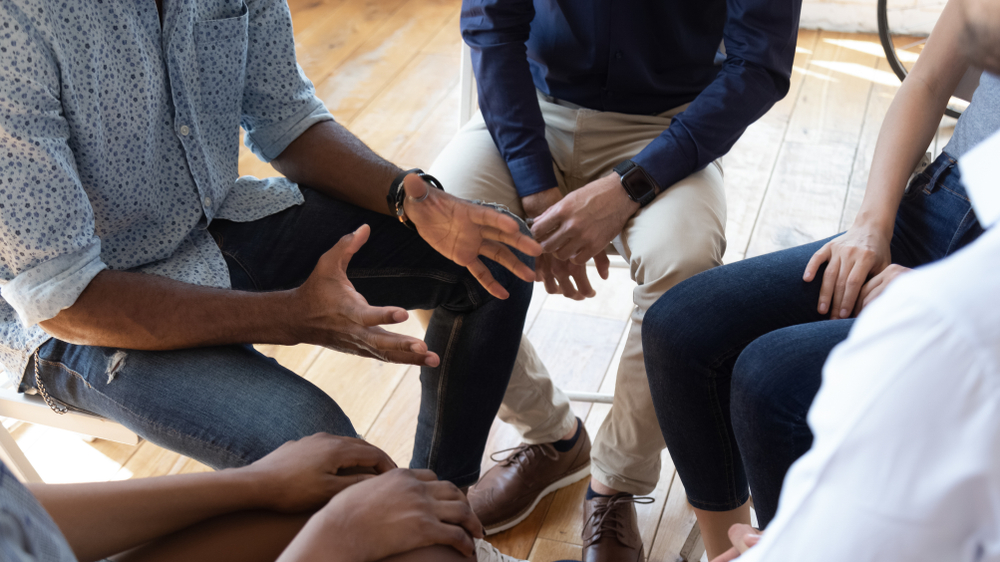 Several individuals in a group setting, seen from above. One person is gesturing with open hands while speaking, surrounded by others who are listening attentively. This image represents open communication and mutual support within a group.
