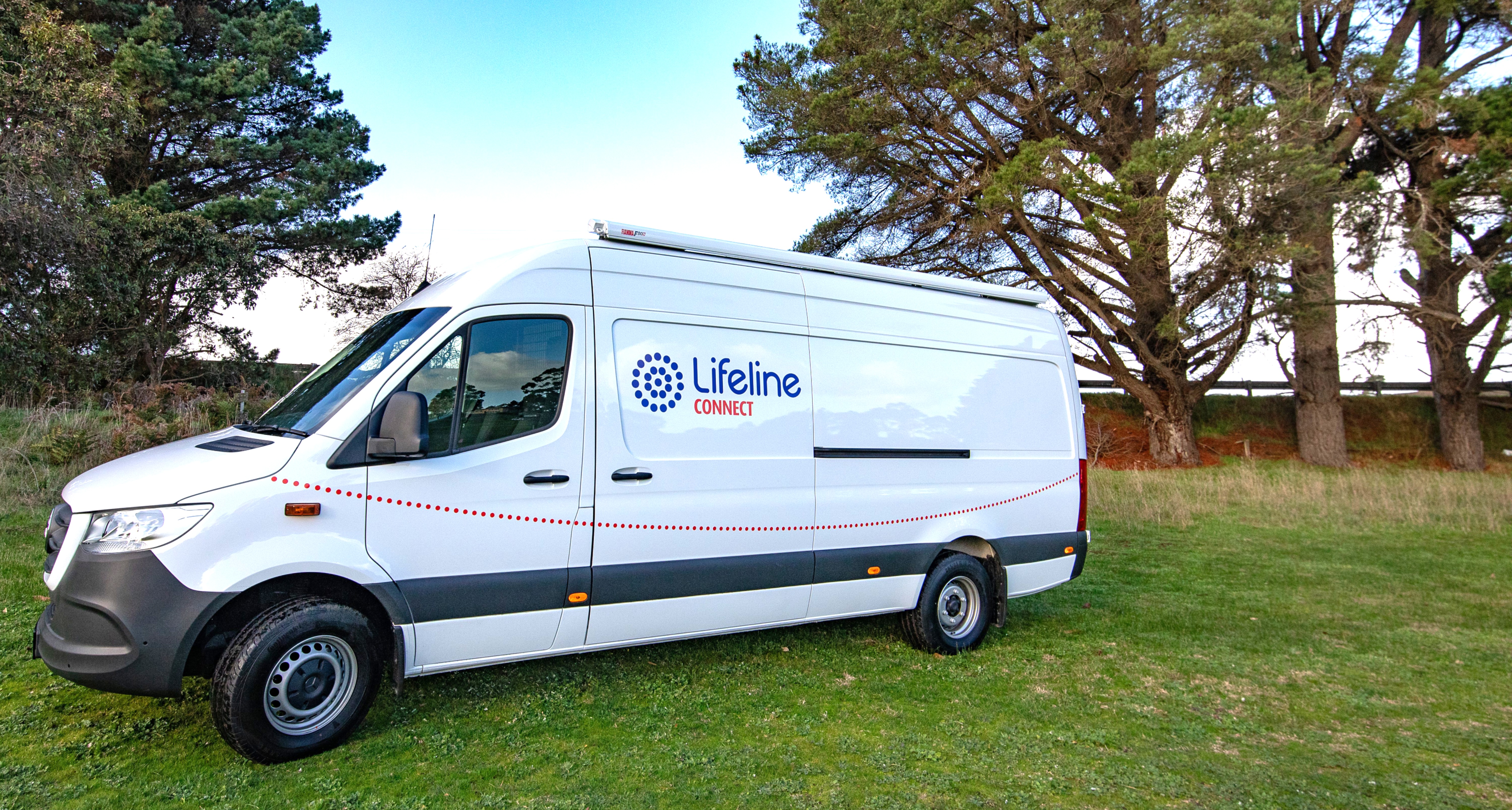 White Lifeline Connect van with blue and red logo, red dotted line graphics, and a dark grey bumper, parked on green grass. Tall pine trees and a fence are visible in the background against a light blue sky.