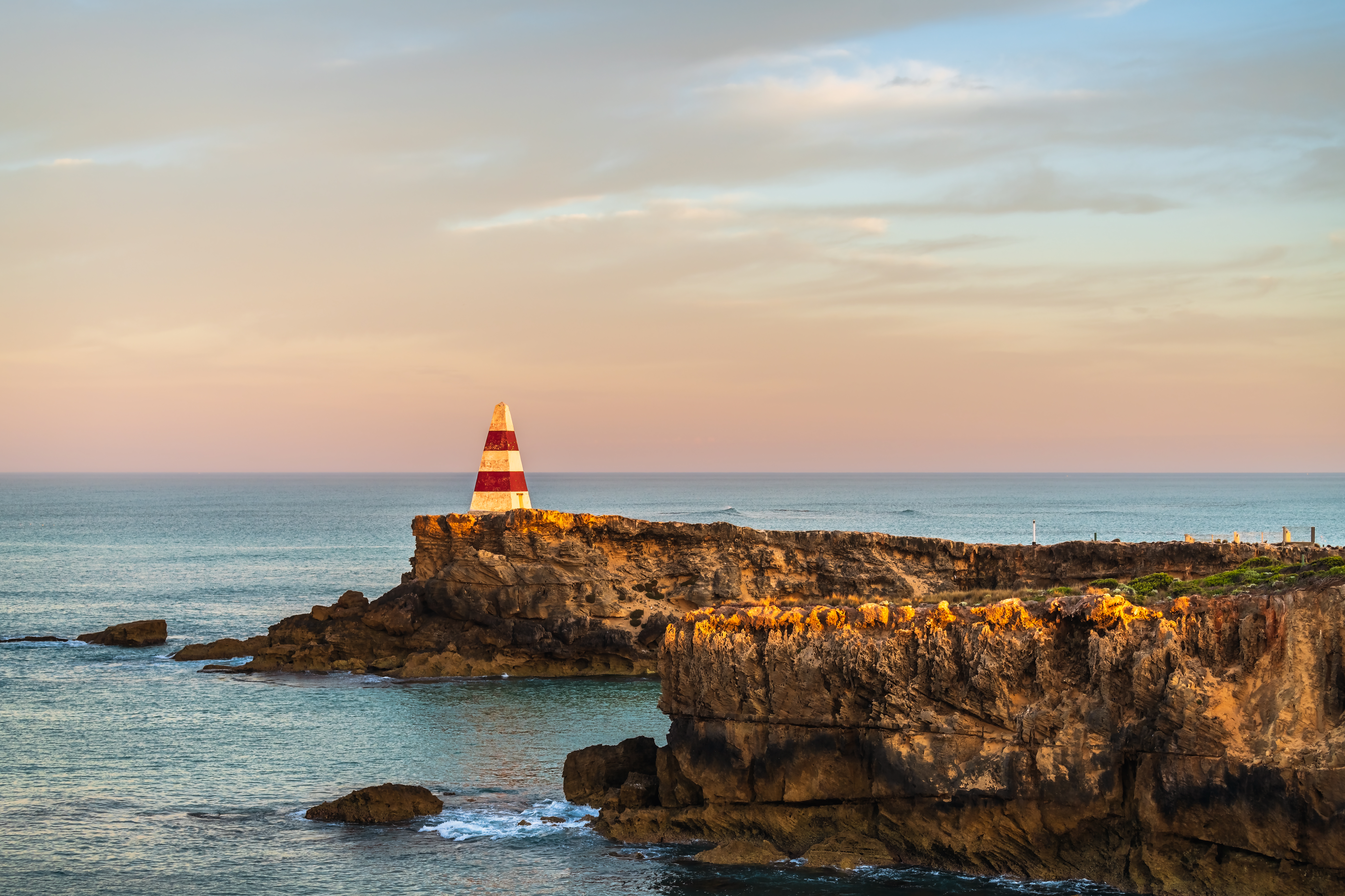 The red and white striped Robe Obelisk stands on a rocky cliff overlooking the calm ocean under a pastel sunset sky. The warm light illuminates the rugged coastline, creating a peaceful and expansive scene.