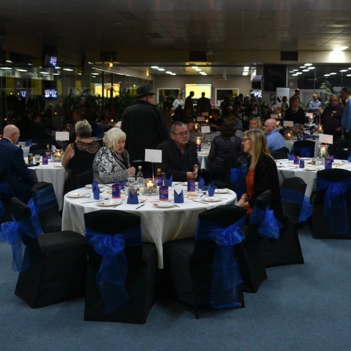 A gala dinner is underway with numerous guests seated at round tables. Tables are draped in white tablecloths, and chairs are covered in black with bright blue sashes. Blue napkins and candles adorn each table. People of various ages are conversing, creating a social atmosphere at the Ballarat and District Suicide Awareness Network Gala.