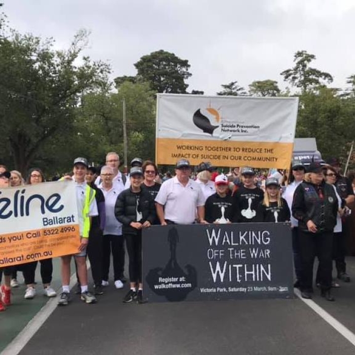 A diverse group of people participating in a suicide prevention event. They hold a Lifeline Ballarat banner with contact details (Call 5322 4996, www.lifelineballarat.com.au) and a sign for 'Walking Off The War Within' with registration info (walkoffww.com) and event date (Victoria Park, Saturday 23 March). A larger banner in the background reads 'Suicide Prevention Network Inc. Working Together to Reduce the Impact of Suicide in Our Community'.