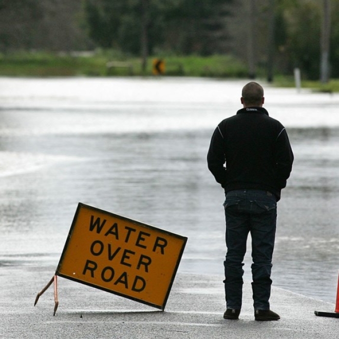 A man standing on a road in front of floodwaters, looking away from the camera. A sign reads "WATER OVER ROAD" to his left, to warn drivers.