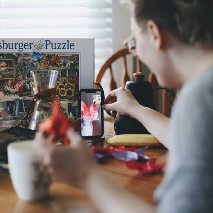 A person sitting at a wooden table doing a video call, showing colorful paper crafts to someone on their phone.