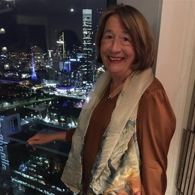 A smiling woman in a brown shirt and light scarf stands in front of a large window, looking out at a vibrant city skyline illuminated at night. Tall buildings with many lights stretch into the distance, and the reflections of the city lights shimmer on water below. Two empty wine glasses sit on a dark surface in the foreground.