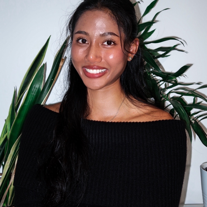 Headshot of a smiling young woman with long dark hair, wearing a black off-the-shoulder top, standing in front of green indoor plants.