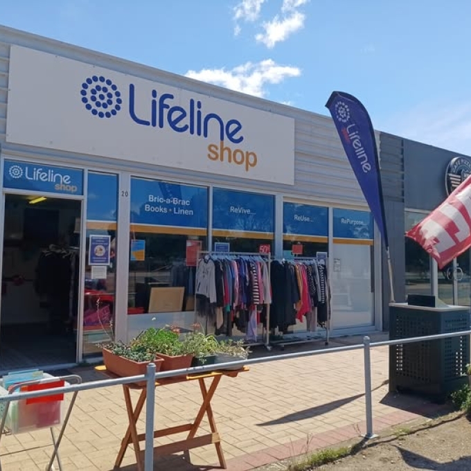 Exterior view of a Lifeline Op Shop with large blue and white signage. A clothing rack displays garments on the paved sidewalk in front, beside a small table with potted succulents. A blue Lifeline flag and a red SALE flag are visible right of the entrance.