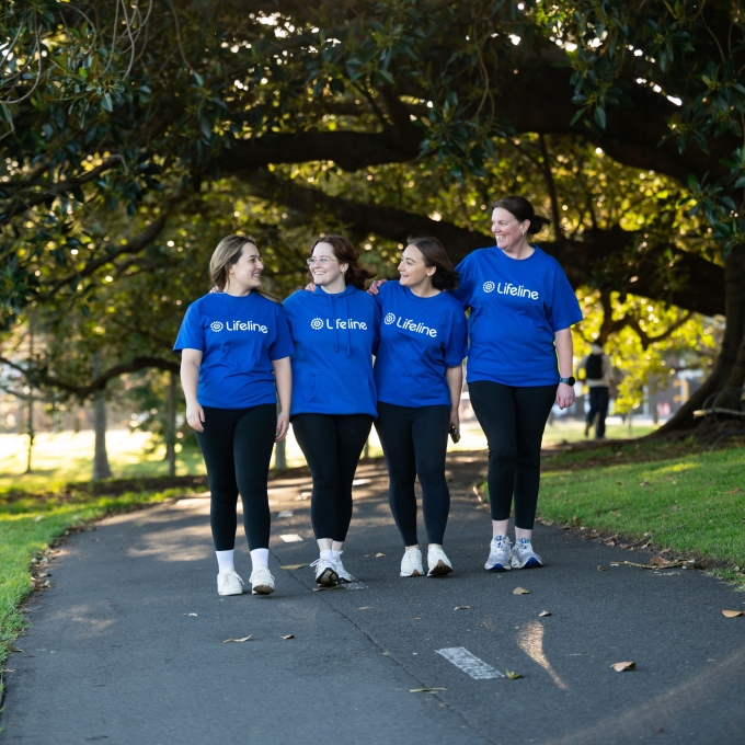 Four women in blue Lifeline t-shirts walk side-by-side down a path in a park, smiling and looking at each other. The sun shines brightly through the leaves of a large tree overhead.