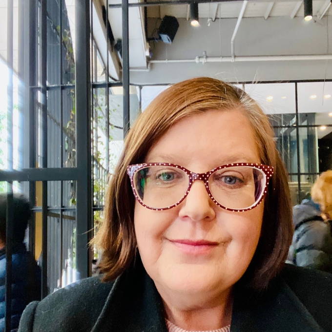 A woman with brown hair and distinctive red-and-white polka dot glasses smiles gently at the camera. She wears a black coat over a light pink sweater, with a soft-focus background of an indoor space featuring large windows and hints of greenery outside.