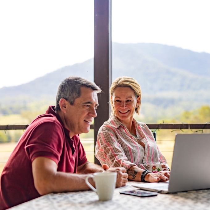 A man and a woman sit at an outdoor table, smiling at each other. The man is on the left, wearing a maroon polo shirt, with a white mug in front of him. The woman, on the right, wears a pink and white striped shirt and is looking at a laptop. In the background, green hills and trees are visible under a bright sky.