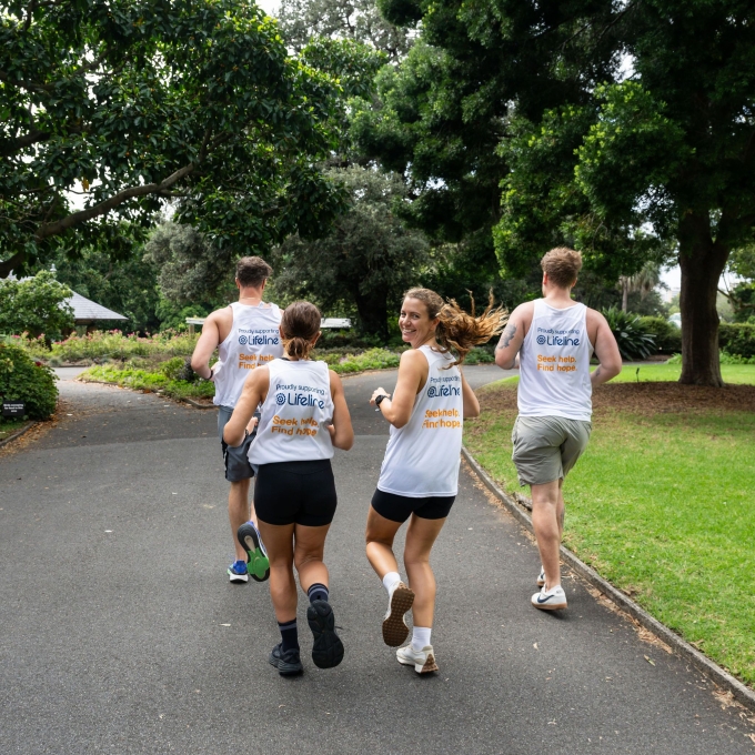 Four people are jogging along a paved path in a lush park. They are seen from behind, but one woman in the middle has turned her head and is smiling at the camera. All four are wearing white singlets with the Lifeline logo and the text "Proudly supporting Lifeline. Seek help. Find hope." The path is bordered by green grass and large, leafy trees. In the background, a white gazebo with a grey roof is partially visible amongst the foliage. The overall impression is one of health, community, and support.