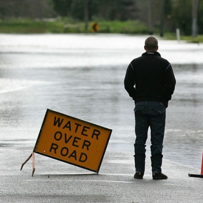A man standing on a road in front of floodwaters, looking away from the camera. A sign reads "WATER OVER ROAD" to his left, to warn drivers.