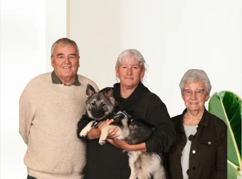 A man, a woman, and an elderly woman are standing together, with the woman in the middle holding a gray and white dog wearing a "Remi" harness. The man on the left wears a cream sweater and green polo. The woman in the middle has gray hair and wears a dark hoodie. The elderly woman on the right has gray hair, glasses, and wears a dark jacket over a light shirt. A green leafy plant is partially visible on the far right.