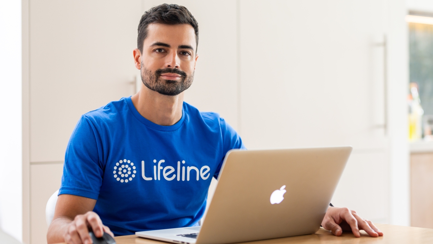 A man with dark hair and a beard, wearing a blue Lifeline t-shirt, sits at a light wooden table. He is holding a computer mouse with his right hand and has a silver laptop open in front of him. He is looking directly at the camera with a calm, friendly expression. The background is a bright, modern kitchen with light-coloured cupboards.